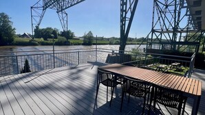 Une terrasse en bois avec une table et des chaises, donnant sur une rivière, avec un pont en arrière-plan.