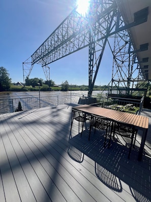Eine Holzterrasse mit Tisch und Stühlen, mit Blick auf einen Fluss und eine Brücke im Hintergrund.