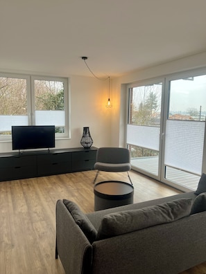 Ein modernes Wohnzimmer mit einem Flachbildfernseher, einem grauen Sofa, einem runden Couchtisch und einer schwarzen Stehlampe.