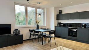 A modern kitchen with dark cabinetry, a dining area with a table and chairs, a television on a stand, and a window with a view of greenery. - Apartment house „Osten – An der Schwebefähre“ – Apartment 4 (Osten)