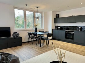 Una cocina moderna con armarios oscuros, un comedor con mesa y sillas, un televisor sobre un mueble bajo y un ventanal con vistas a zonas verdes.