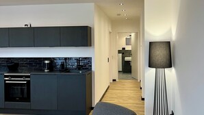 A modern kitchen with dark cabinetry, a built-in stove, and a dining area with a grey sofa and a floor lamp. - Apartment house „Osten – An der Schwebefähre“ – Apartment 4 (Osten)
