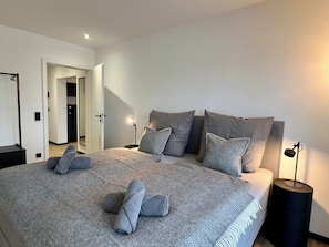 Ein modernes Schlafzimmer mit einem großen Bett, vielen Kissen, Nachttischlampen und einer sichtbaren Tür zu einem anderen Raum.