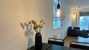 Un'area pranzo moderna con tavolo nero, sedie, un vaso con fiori secchi, un lampadario e vista su un soggiorno con divano e finestra.