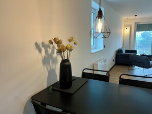 A modern dining area with a black table, chairs, a vase with dried flowers, a hanging light fixture, and a view of a living room with a sofa and a window. - Apartment house „Osten – An der Schwebefähre“ – Apartment 8 (Osten)