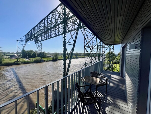 Una terraza con sillas que da a un río, una estructura de puente metálico y una casa con techo inclinado.