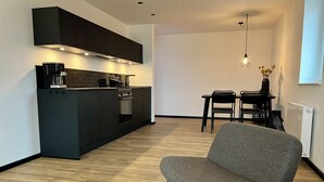 Eine moderne Küche mit dunklen Schränken, einem eingebauten Backofen und einer Kaffeemaschine. Es gibt einen Essbereich mit Tisch und Stühlen, und ein grauer Stoffstuhl im Vordergrund.