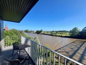 A balcony with a view of a river, a church, and green fields. - Apartment house „Osten – An der Schwebefähre“ – Apartment 10 (Osten)