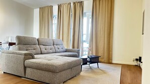 A modern living room with a sectional sofa, a small side table, and a large window with curtains. - Stylish 2BR With 1LR Near to City Center (Ghent)