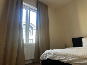 Quarto com cama, janela com cortinas, radiador e vista para um edifício.