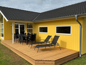 A yellow building with a wooden deck, a dining table with chairs, and two windows. - 4 Star Holiday Home in Grossenbrode (Grossenbrode)