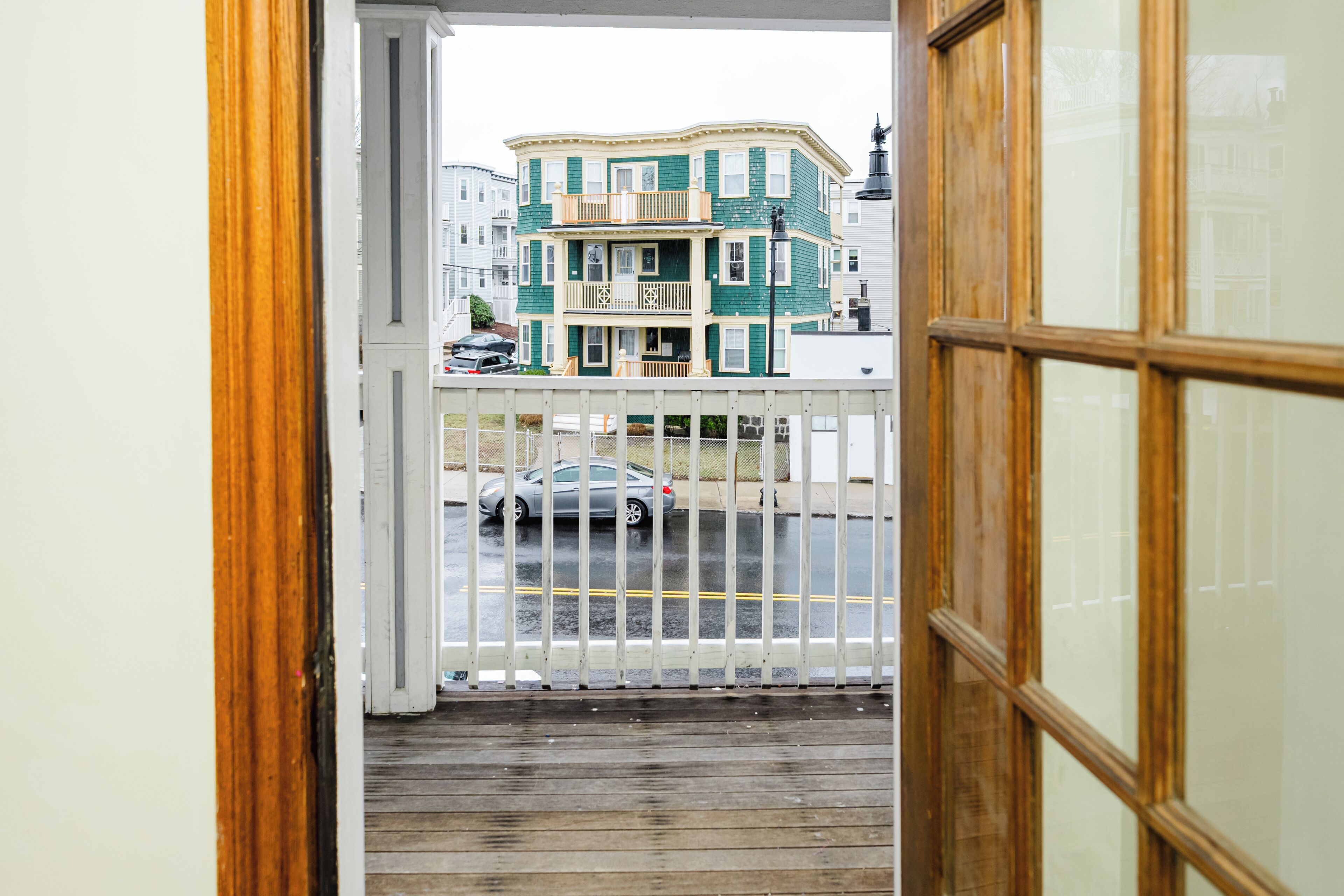 Vista desde un balcón con baranda de madera, una calle con coches y un edificio de varios pisos con fachada verde.