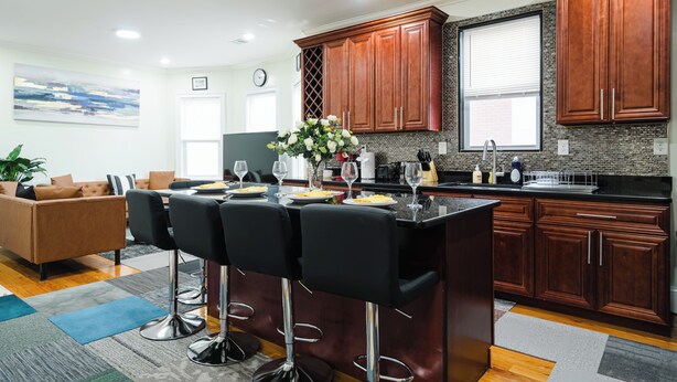 A modern kitchen with dark wooden cabinets, a central island with bar stools, and a dining area with a brown leather sofa. - Spacious & Cozy 4BR Boston Apt with view / Family Gathering - 90/2 (Boston)