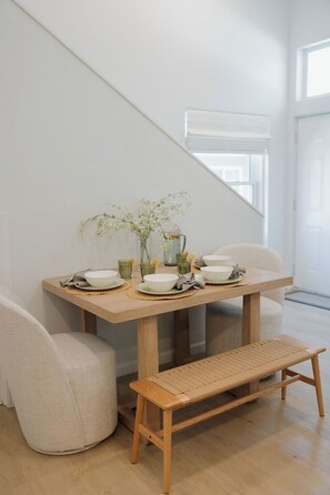 Un coin repas avec une table en bois, des chaises blanches et un banc en osier.