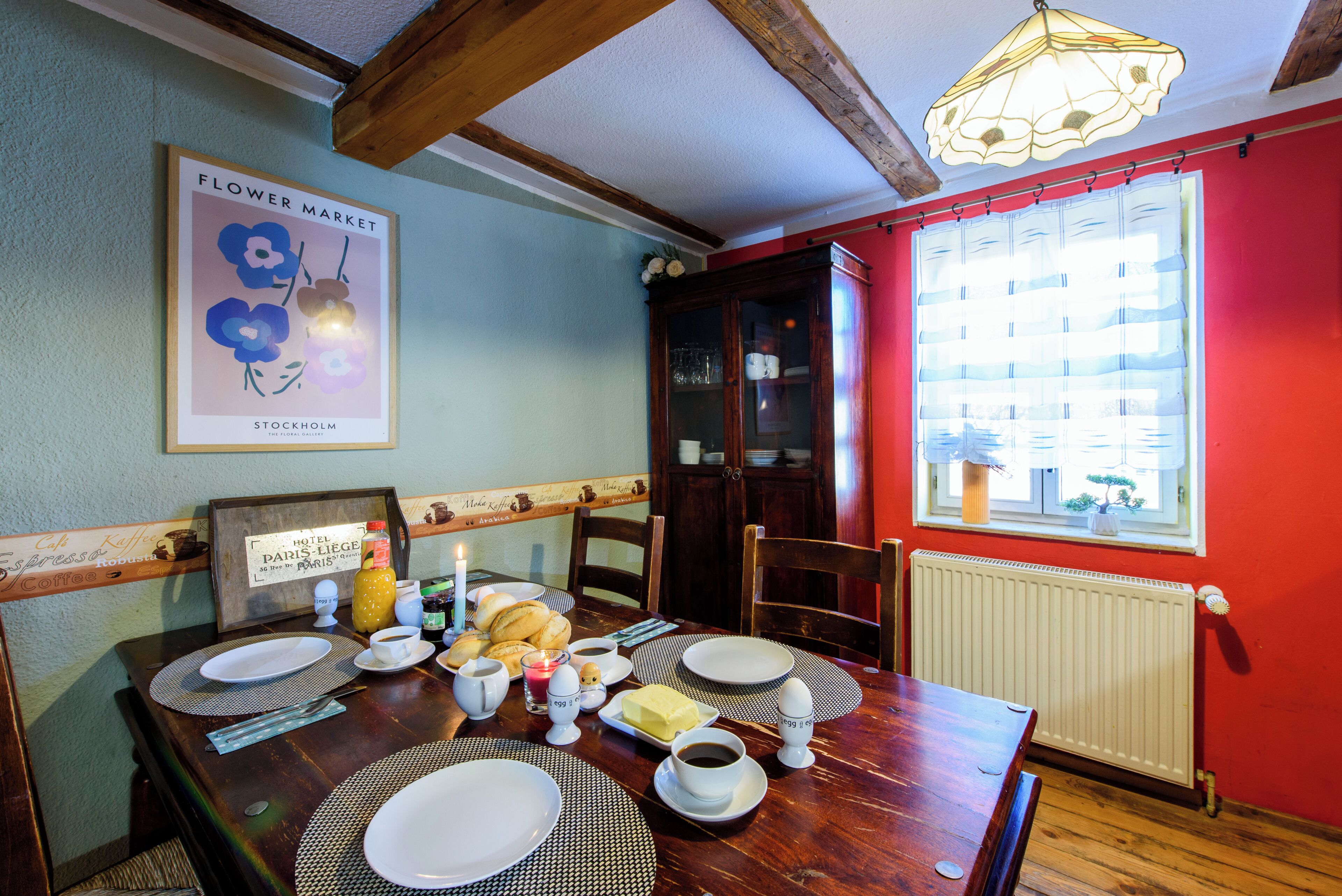 Une table à manger dressée avec des assiettes, des tasses et une bougie, agrémentée d’un cadre décoratif et d’une fenêtre avec des stores.
