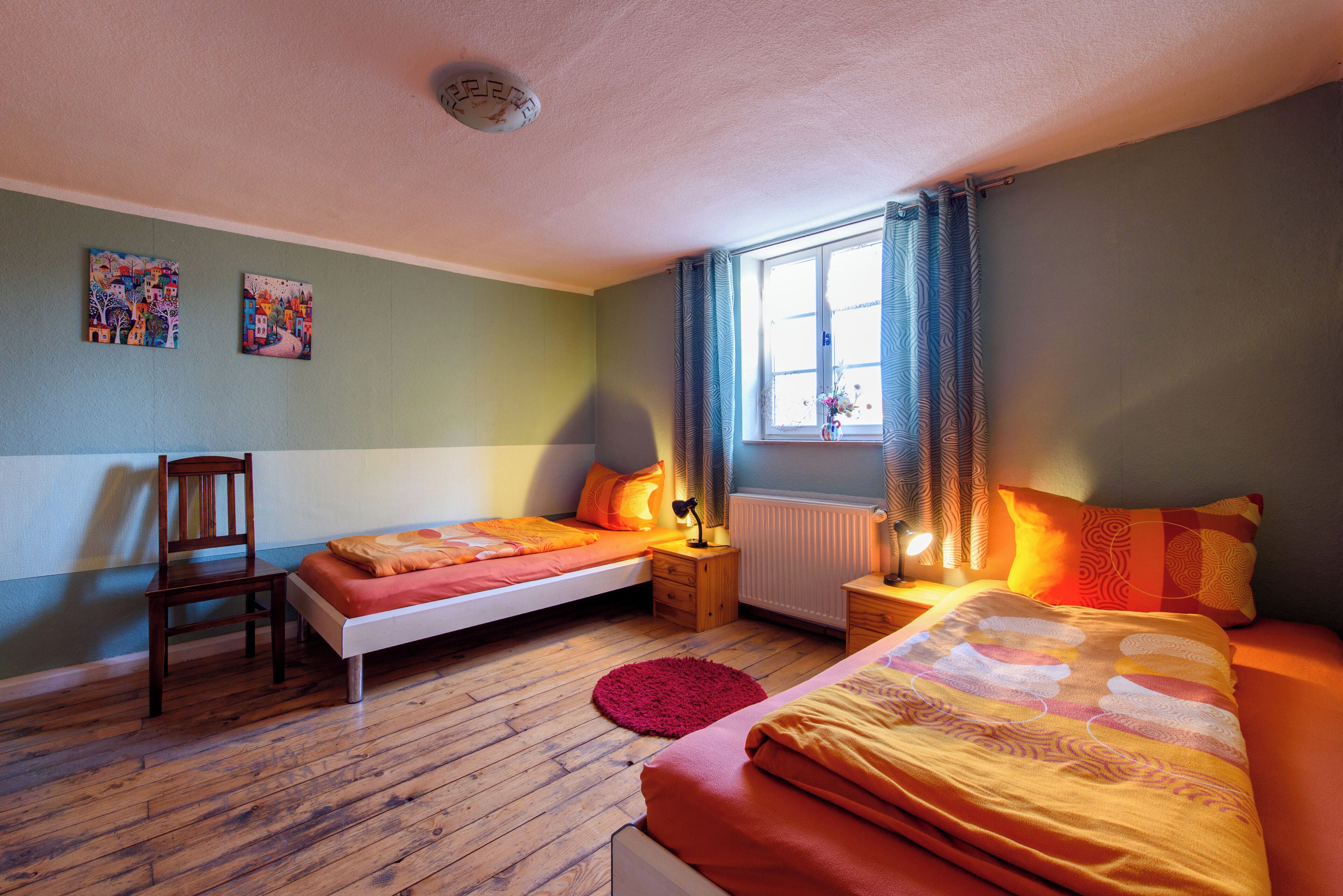 Une chambre avec deux lits, une chaise, une fenêtre avec des rideaux et des œuvres d’art accrochées au mur.