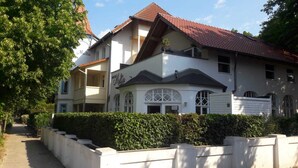 Un bâtiment à deux étages, avec un toit recouvert de tuiles rouges, un balcon à l’étage supérieur et une haie devant.