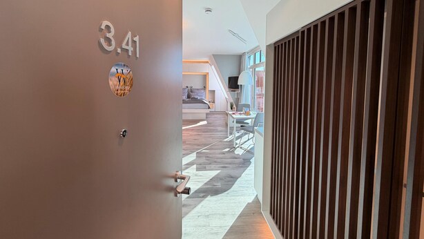 A modern hotel corridor with a door labeled '3.41' and a view into a room with a bed, desk, and window. - Anna Düne 3-41 - Dream location in the Aparthotel Anna Düne with a view of the Wadden Sea (Wangerooge)