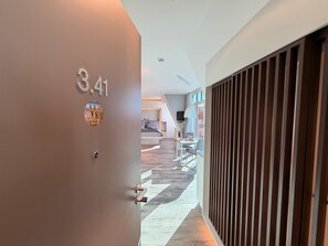 En modern hotellkorridor med en dörr märkt med siffran ’3.41’ och en utsikt in i ett rum med en säng, ett skrivbord och ett fönster.