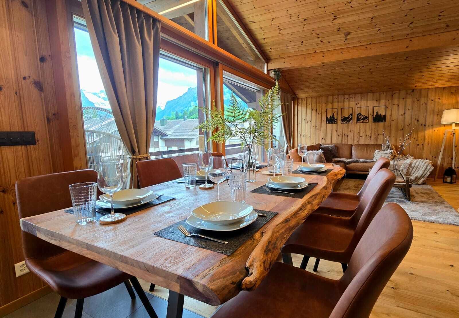 Ein Holztisch ist gedeckt mit Tellern, Gläsern und Besteck, umgeben von Ledersesseln, mit Blick auf Berge durch große Fenster.