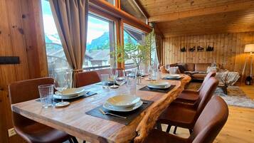 Ein Holztisch ist gedeckt mit Tellern, Gläsern und Besteck, umgeben von Ledersesseln, mit Blick auf Berge durch große Fenster.