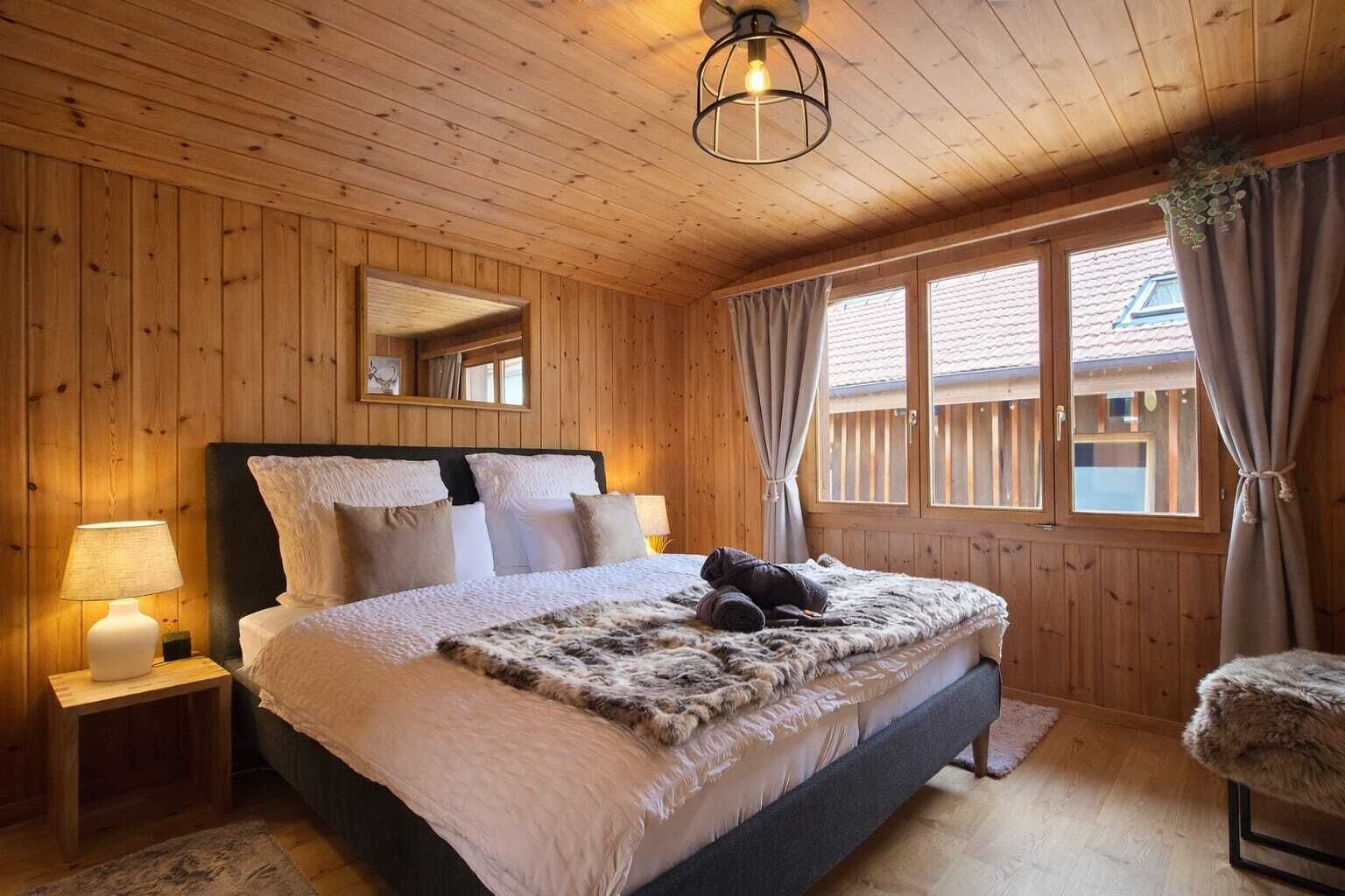 Quarto com painéis de madeira, uma cama, mesas de cabeceira, um espelho e uma janela com cortinas.