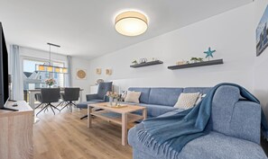 A modern living room with a blue sofa, wooden coffee table, and a dining area with a table and chairs. - Ostseesternchen - Sauna, washing machine, terrace, private parking space (Olpenitz)
