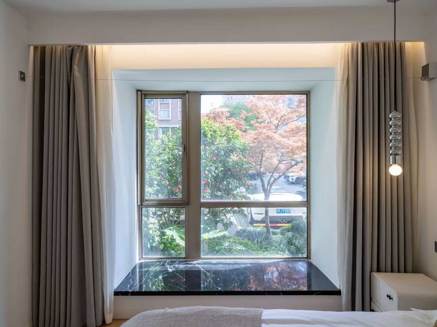 Una habitación de hotel con un ventanal grande, una cama, una mesita de noche y una lámpara.
