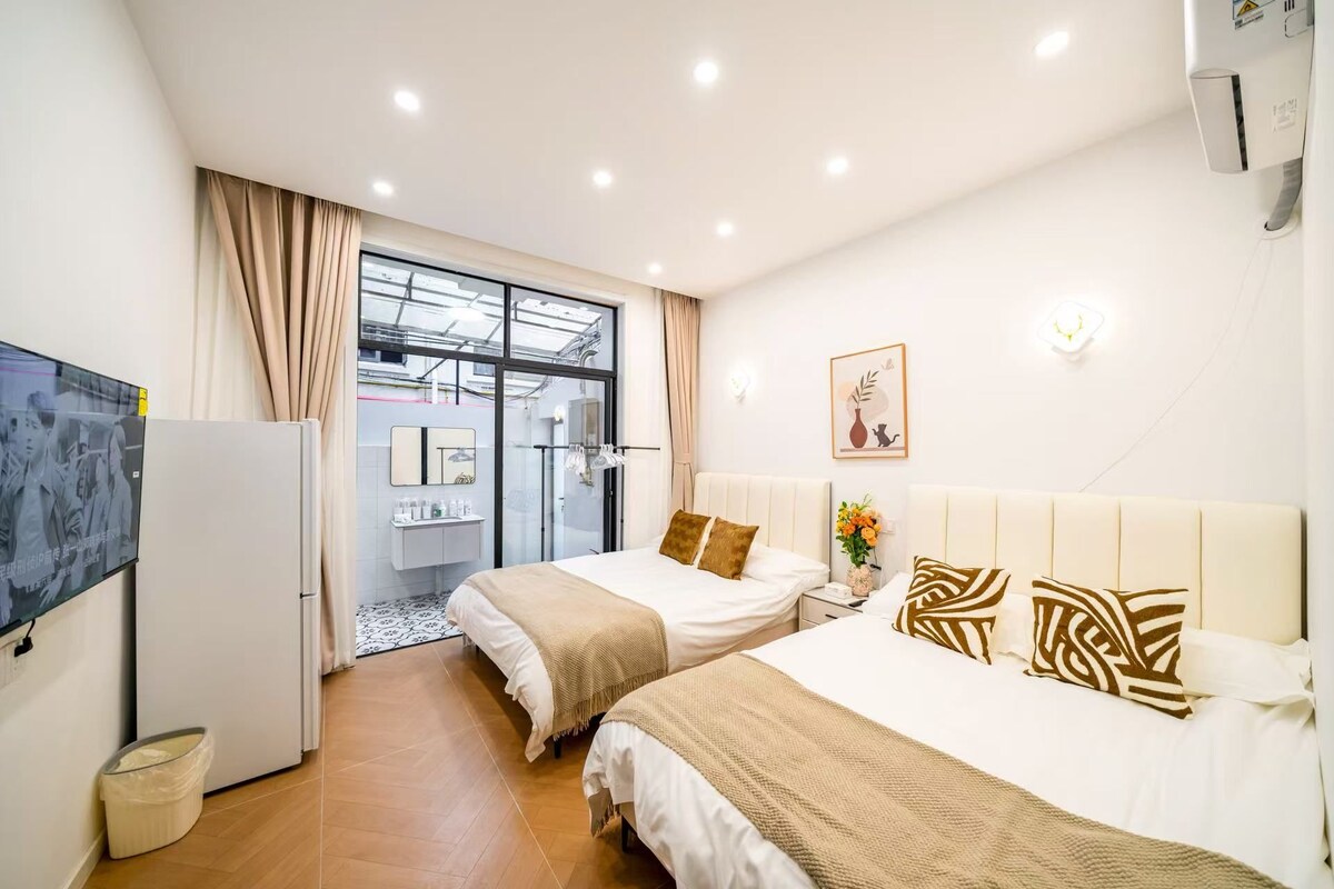 A hotel room with two beds, a television, a mirror, a small table with a vase, and a window with curtains.