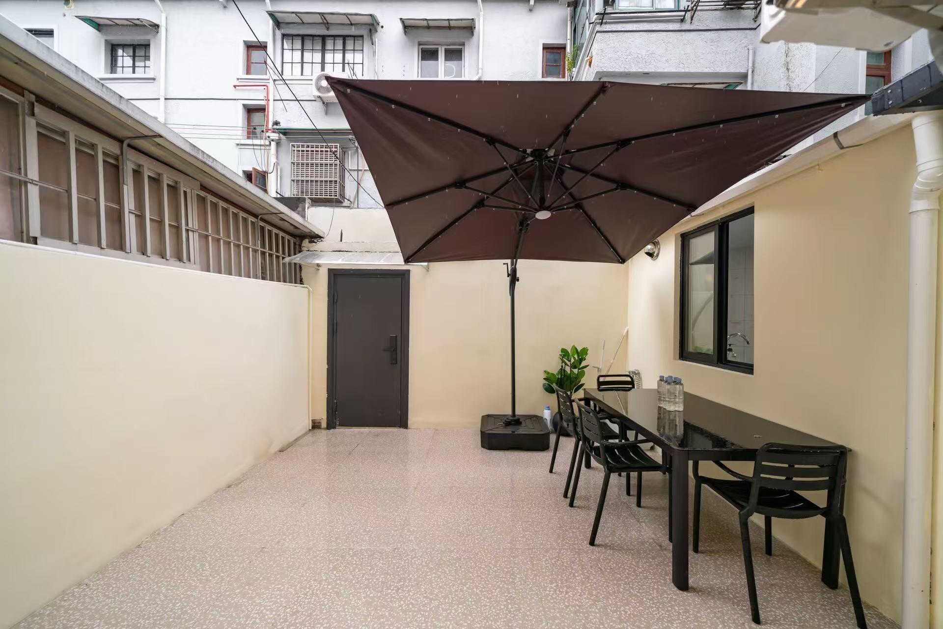 A narrow outdoor patio with a table and chairs, a large umbrella, and a potted plant.