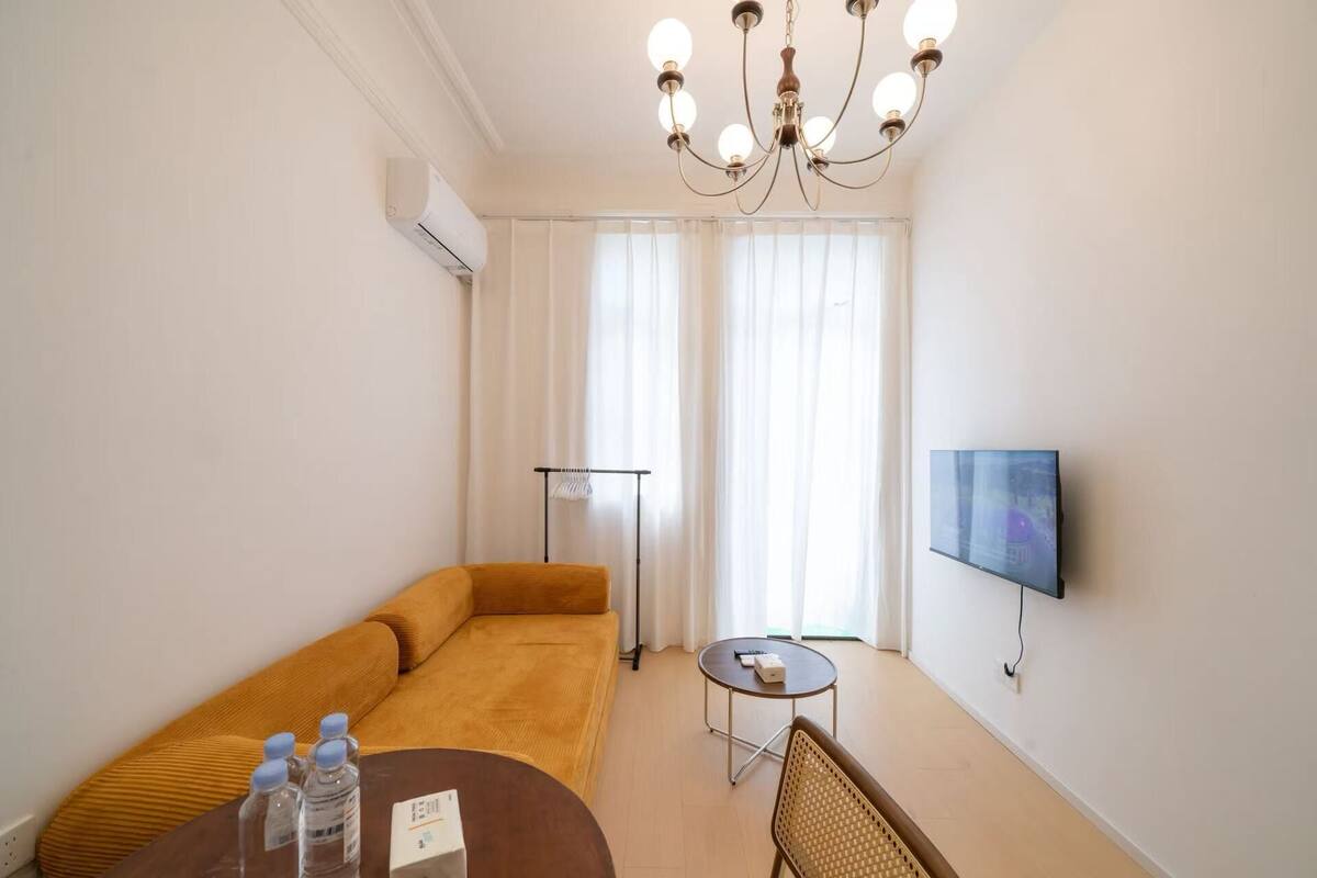 Moderni hotellihuone, jossa on sohva, pieni pyöreä pöytä, jossa on kaukosäädin, seinälle asennettu litteänäyttöinen televisio ja lattiavalaisin.