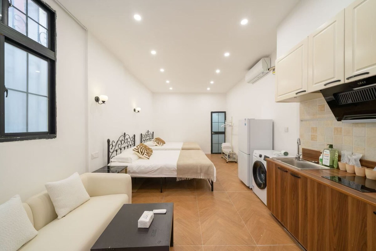 A compact hotel room with a bed, a sofa, a small table, a washing machine, and a kitchenette.