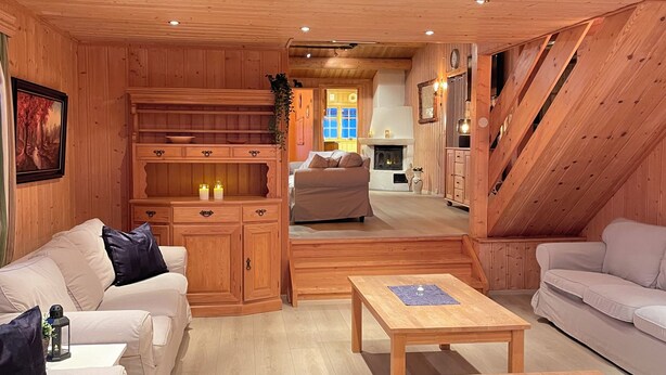 Ein gemütliches Blockhaus-Interieur mit Holzwänden, Kamin, einer Couch, einem Esstisch und einer Treppe.