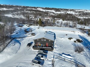 A large building with a flat roof, surrounded by a parking area with several cars, set in a snowy landscape with trees and distant buildings. - Large cabin 27 guests with panoramic view (Buskerud)