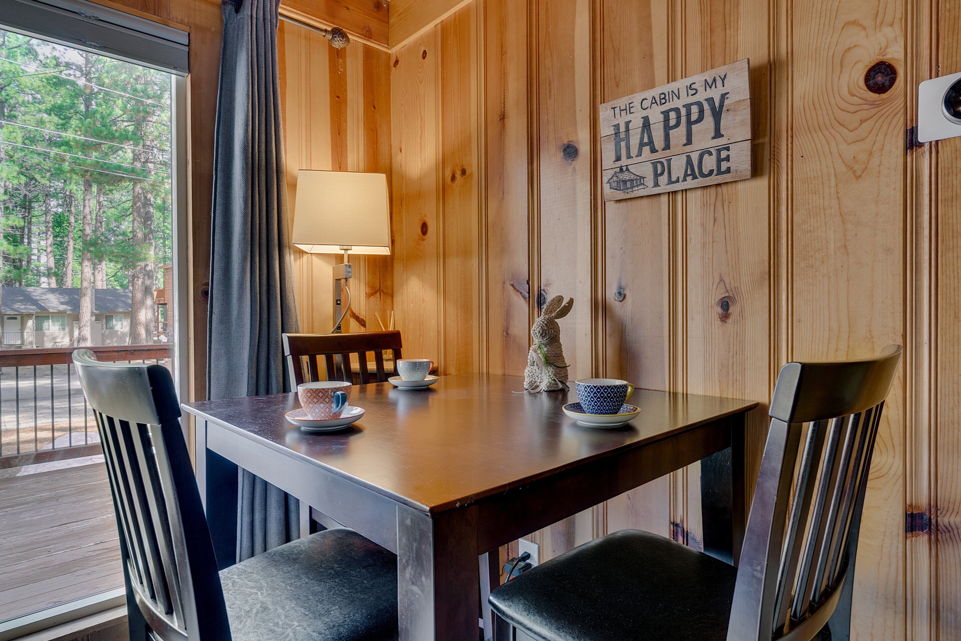Interior de una cabaña de madera con una mesa de comedor, sillas, una lámpara y un cartel que dice 'The cabin is my happy place'.