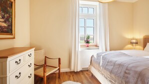 Um quarto com uma cama, cômoda, cadeira, janela com cortinas e luminária.