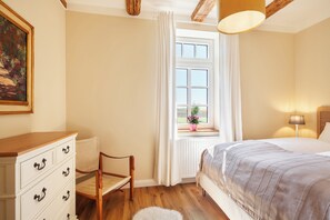 A bedroom with a bed, a dresser, a chair, a window with curtains, and a lamp. - Vacation home Kreuzkate near Grömitz - Baltic Sea & fireplace (Manhagen)