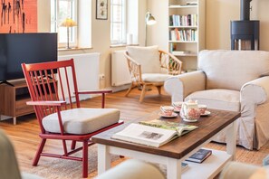 Ein gemütliches Wohnzimmer mit einem roten Sessel, einer Couch, einem Holztisch mit Büchern und Teetassen sowie einem Bücherregal im Hintergrund.