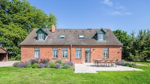 Uma casa de dois andares de tijolo com varanda frontal, uma pequena garagem anexa e um jardim bem cuidado com diversas plantas e flores.