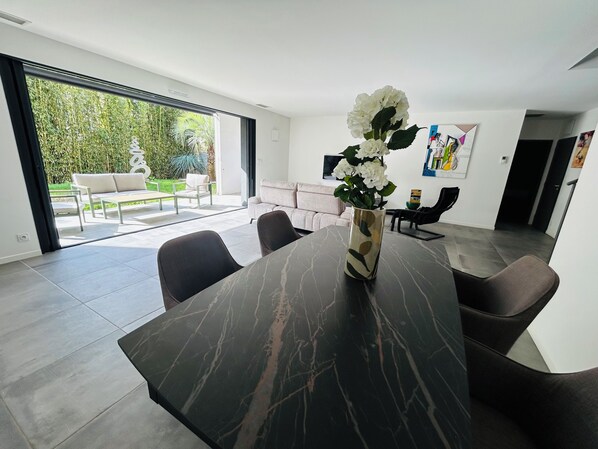 Un espace repas moderne avec une table en marbre, un vase rempli de fleurs blanches et une vue sur un jardin, visible à travers des portes vitrées.
