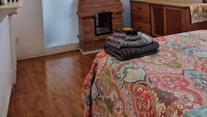 Une chambre avec une cheminée en briques, un lit recouvert d’une courtepointe colorée, une commode en bois et un tableau accroché au mur.