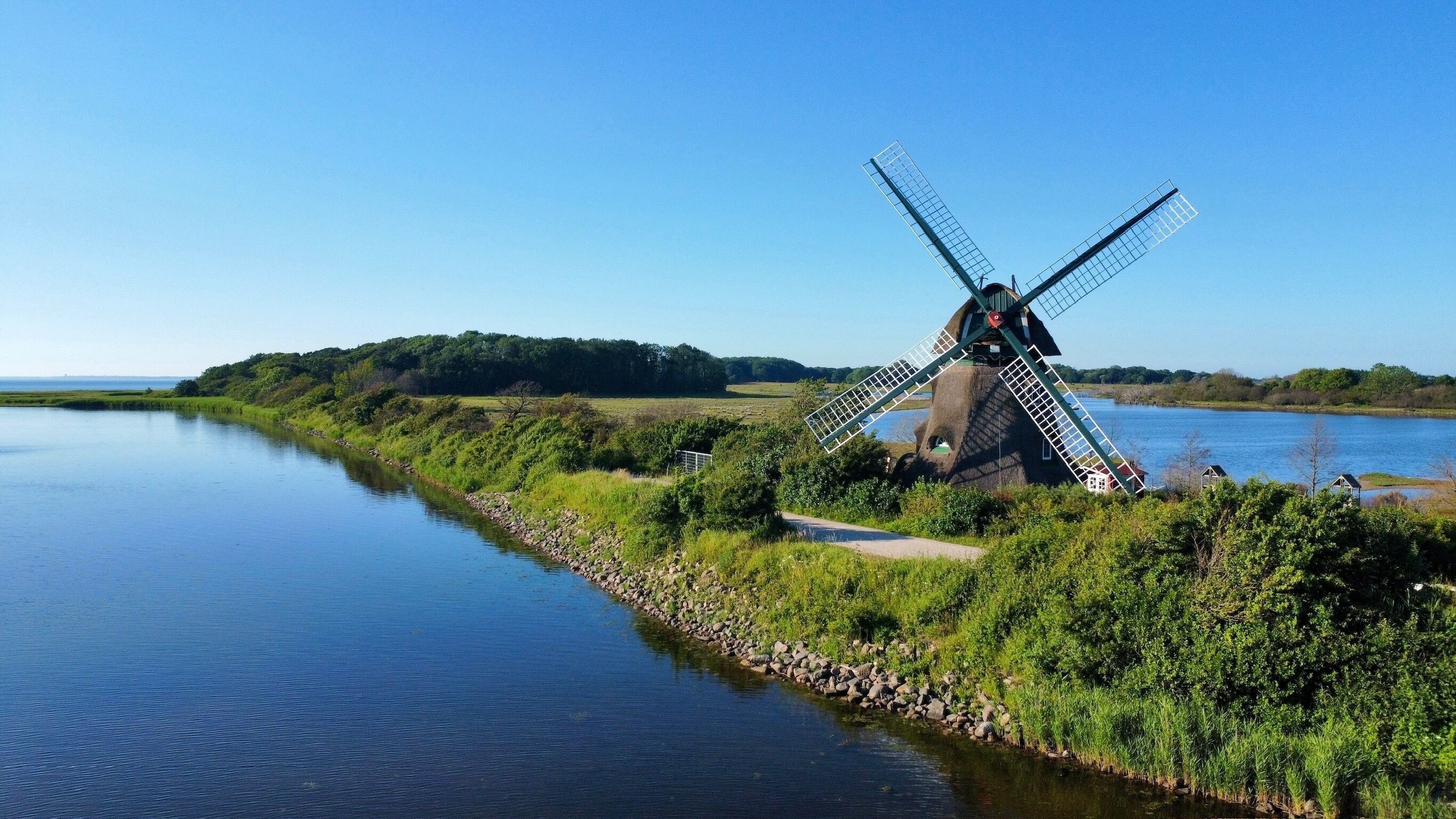 A windmill by a calm river, surrounded by greenery and a clear sky.