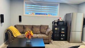A living room with a brown sofa, a coffee table, a black cabinet with glass doors, a stainless steel refrigerator, and a window with blinds. - Entire Basement suite (Chestermere)