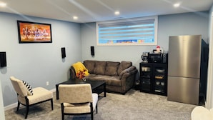 A living room with a sofa, two chairs, a coffee table, a refrigerator, a microwave, a window with blinds, a painting, and a speaker. - Entire Basement suite (Chestermere)
