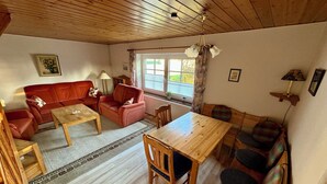 Uma sala de estar aconchegante com teto de madeira, um sofá vermelho, uma mesa de centro de madeira, uma mesa de jantar com cadeiras e uma janela com cortinas.