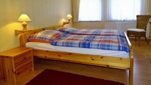 Uma cama de madeira com colcha xadrez, duas luminárias de cabeceira, uma mesa de cabeceira de madeira, uma cadeira de vime e uma janela com cortinas finas.