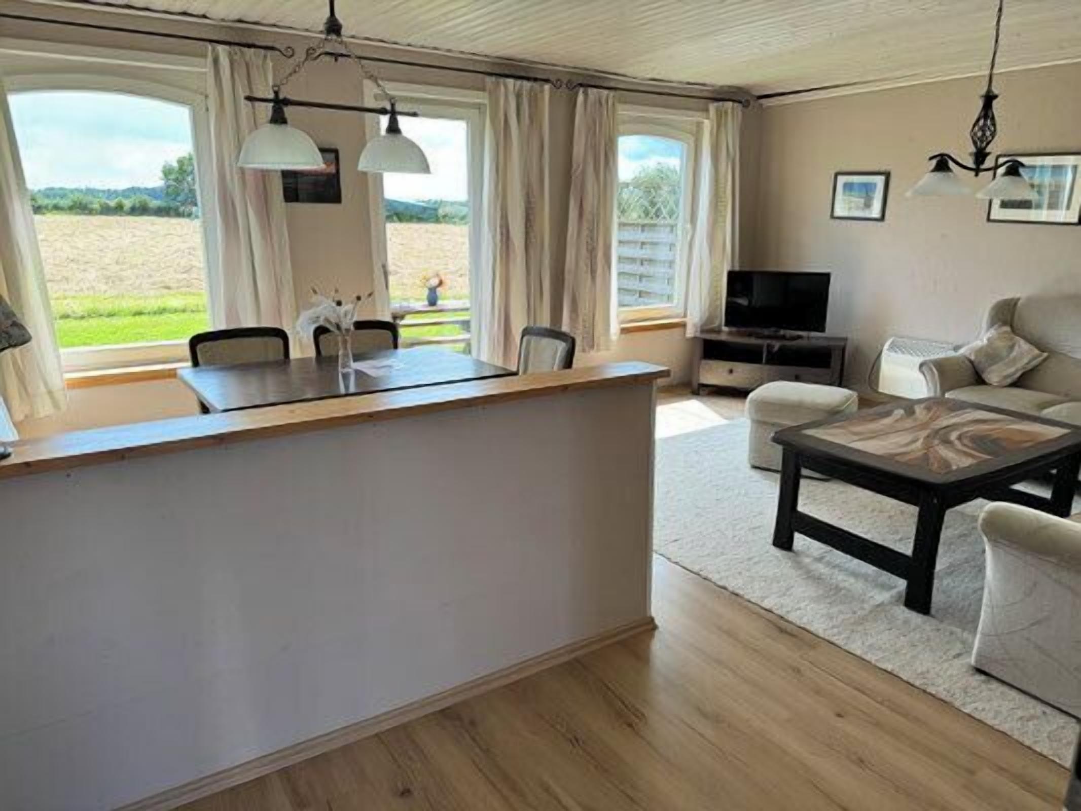 Amplia sala de estar con mesa de comedor de madera, sofá, televisor y ventanas grandes con vistas al exterior.