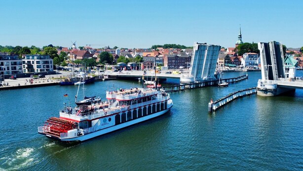 Un río con un barco grande amarrado a un muelle, un puente y edificios al fondo.