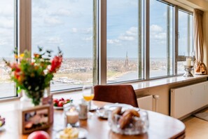 A dining area with a table set for breakfast, a vase of flowers, and a view of a cityscape through large windows. - Vacation apartment "FjordLoft" on the 24th floor of the Viking Tower (Schleswig)