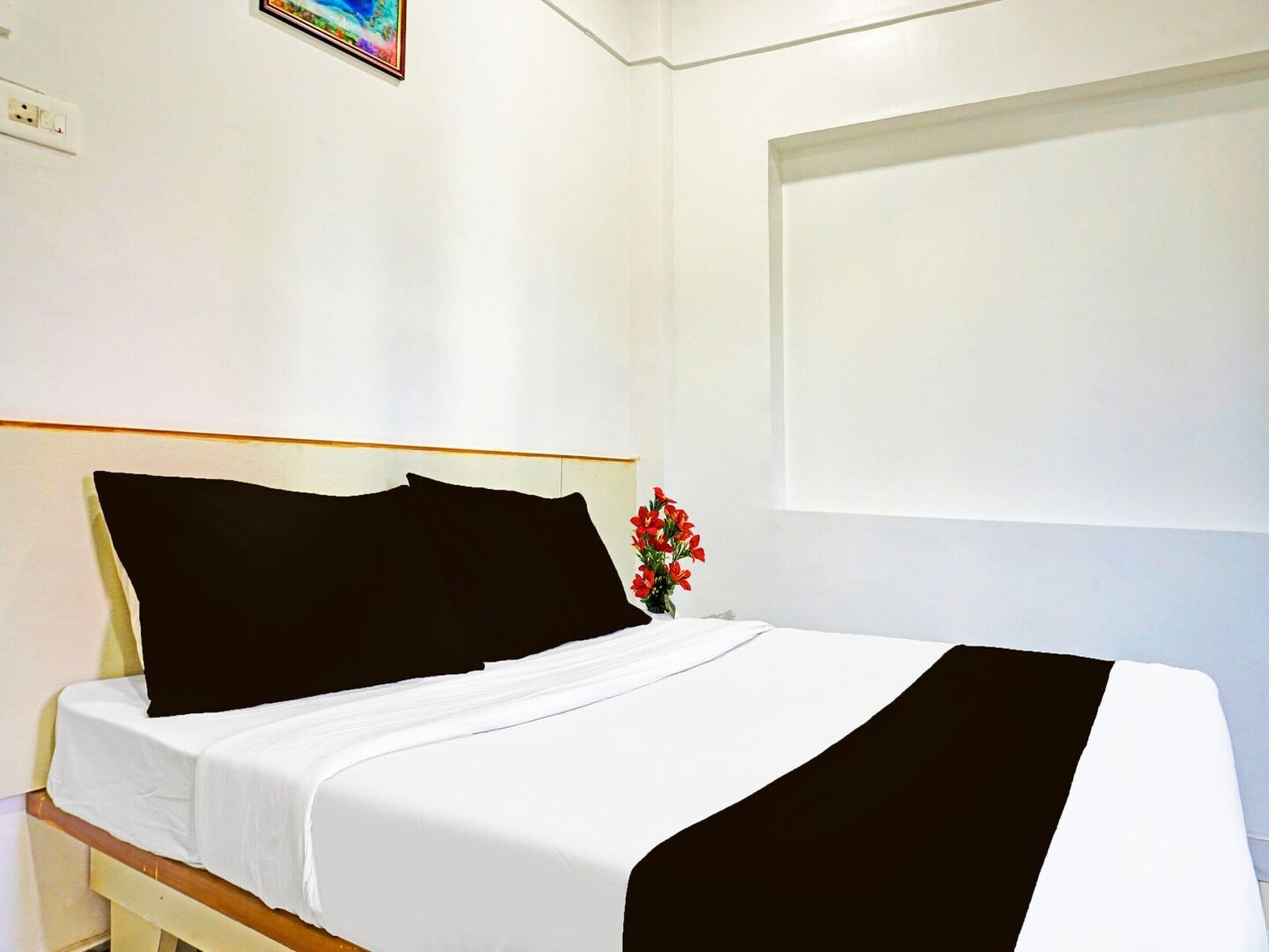 Ein Bett mit weißer Bettwäsche und schwarzen Kissen, eine kleine Vase mit roten Blumen und eine weiße Wand mit einer Steckdose.